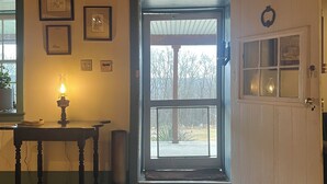 A room with a wooden ceiling, a door leading outside, a table with a lamp, and framed pictures on the wall.