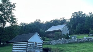 A rustic cabin with a stone foundation and a wooden exterior, surrounded by a stone wall and a grassy field.
