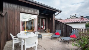 Edificio de madera con un patio que incluye una mesa redonda, sillas blancas y dos sillones con cojines rojos.