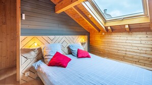 Un dormitorio con techo inclinado, paredes de madera, cama con ropa de cama azul y almohadas rojas, y un tragaluz.
