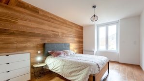A bedroom with a wooden wall, a bed with a headboard, a bedside table with a lamp, and a window providing a view. - Ouréa - duplex avec balcon (La Bresse)