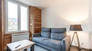 A modern living room with a grey sofa, a small table, a floor lamp, and a window with a view of buildings and hills. - Ouréa - duplex avec balcon (La Bresse)