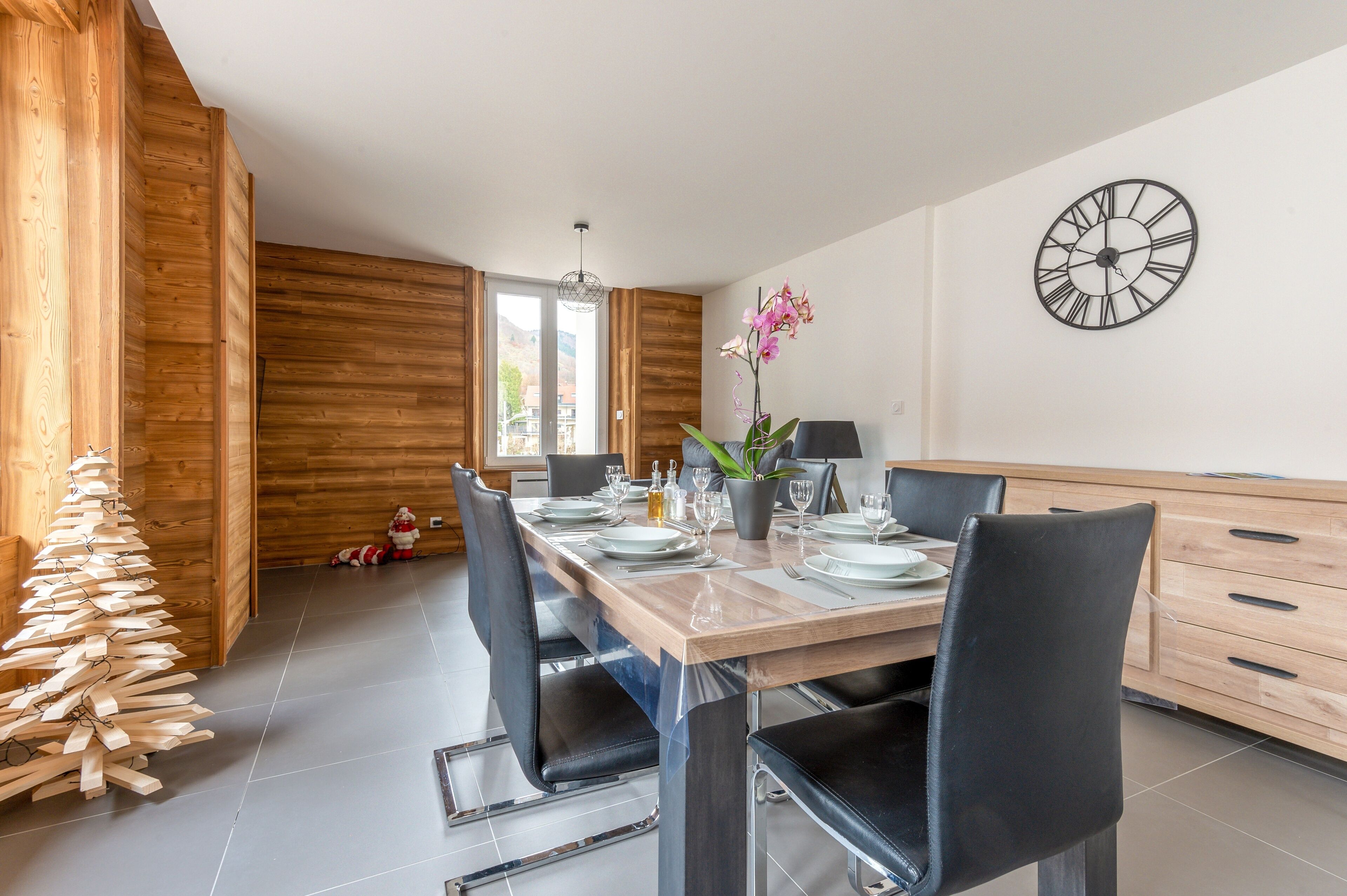 A modern dining area with a wooden table set for four, black chairs, a decorative plant, and a wall clock.