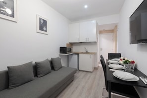 A modern, compact living space with a grey sofa, a dining table set for two, a microwave, a small refrigerator, and a flat-screen TV mounted on the wall.