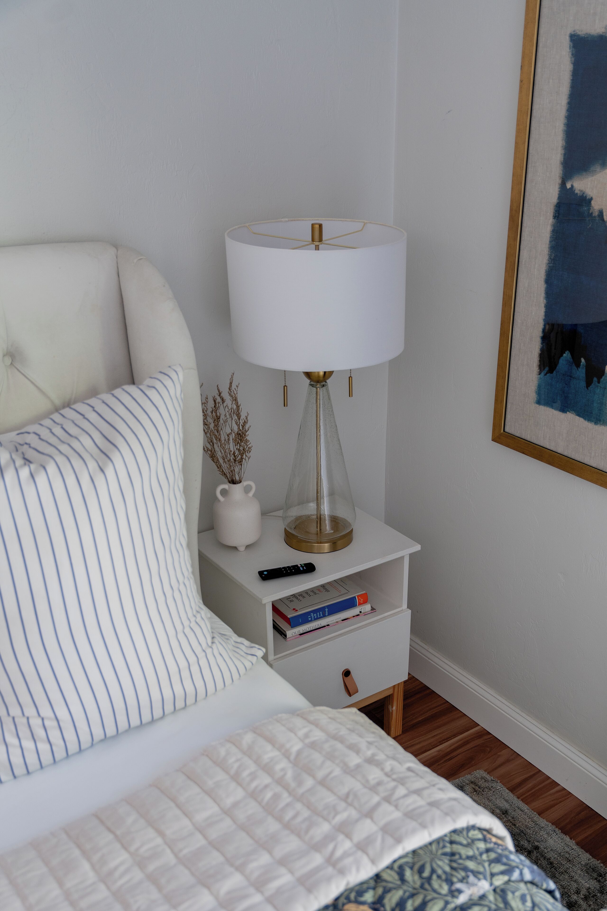 A neatly made bed with a striped pillow and a quilted blanket, a bedside table with a lamp, a vase with dried plants, and a framed picture on the wall.