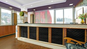 Zona de recepción de un hotel con un mostrador de madera, un ventanal con vistas a edificios y un tablón de anuncios.