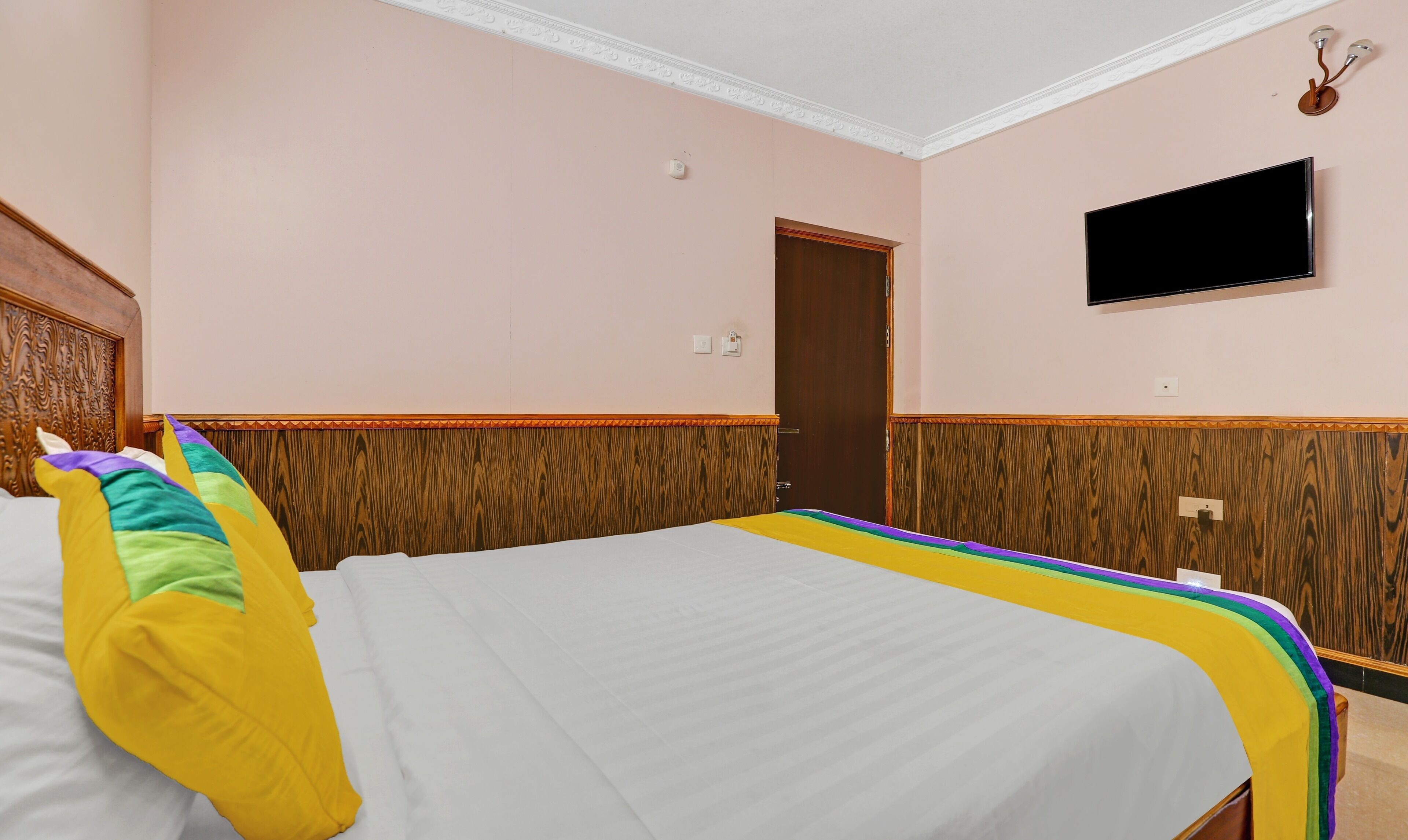 A hotel room with a large bed, a wooden headboard, a television mounted on the wall, and a door.