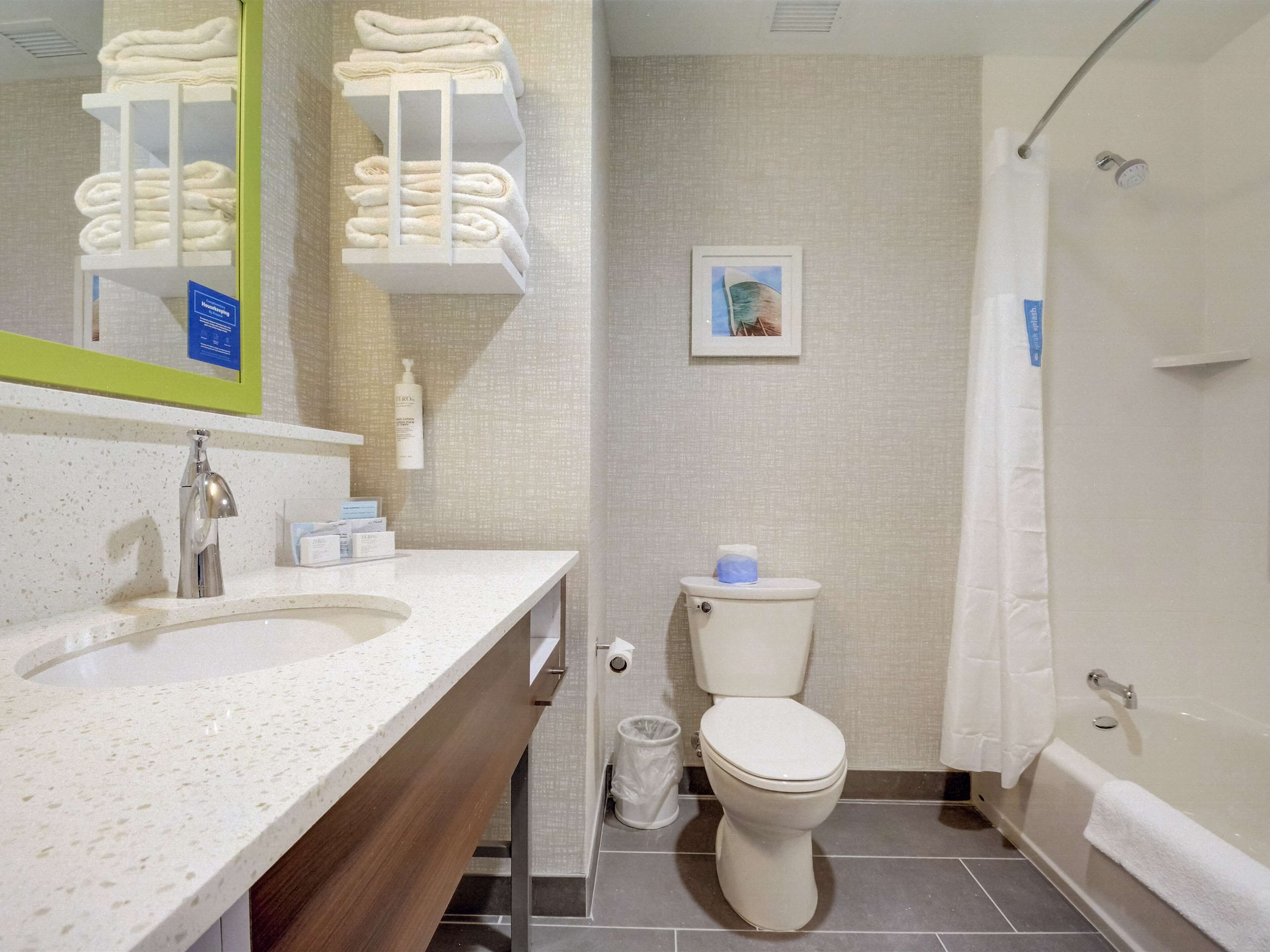 Een badkamer met een wastafel, toilet en bad. Er is een handdoekhouder met gevouwen handdoeken, een foto aan de muur en een prullenbak.
