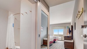 A hotel room with a large mirror, a bed with two pillows, a bedside table, a window with a view, and a shower area.