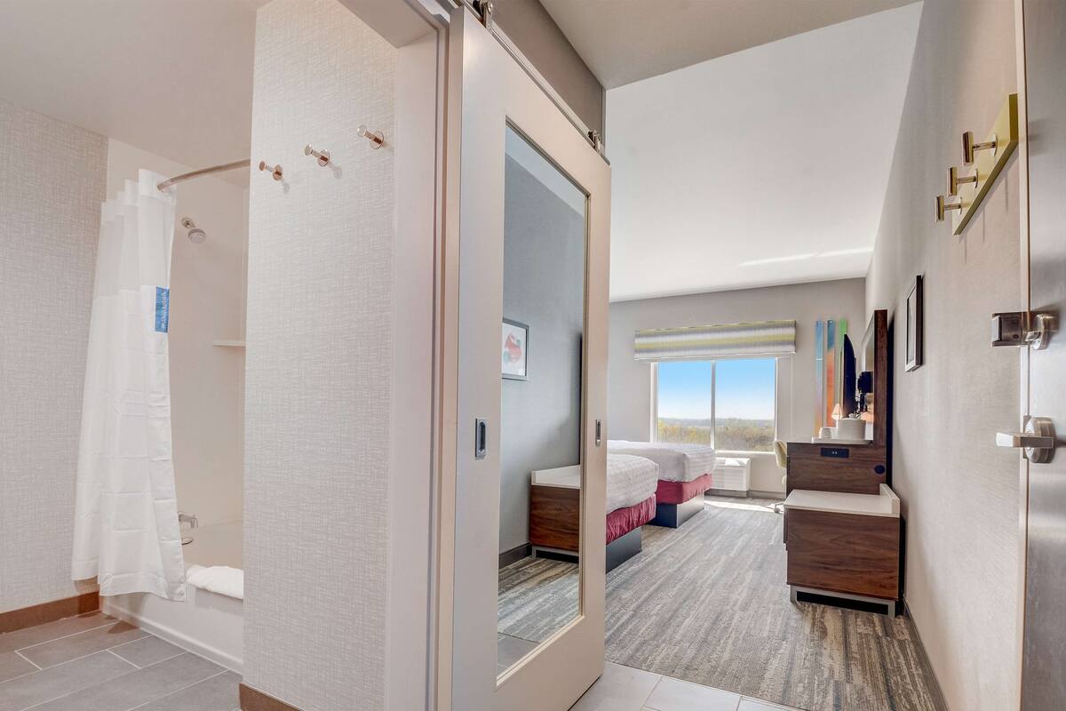 A hotel room with a large mirror, a bed with two pillows, a bedside table, a window with a view, and a shower area.