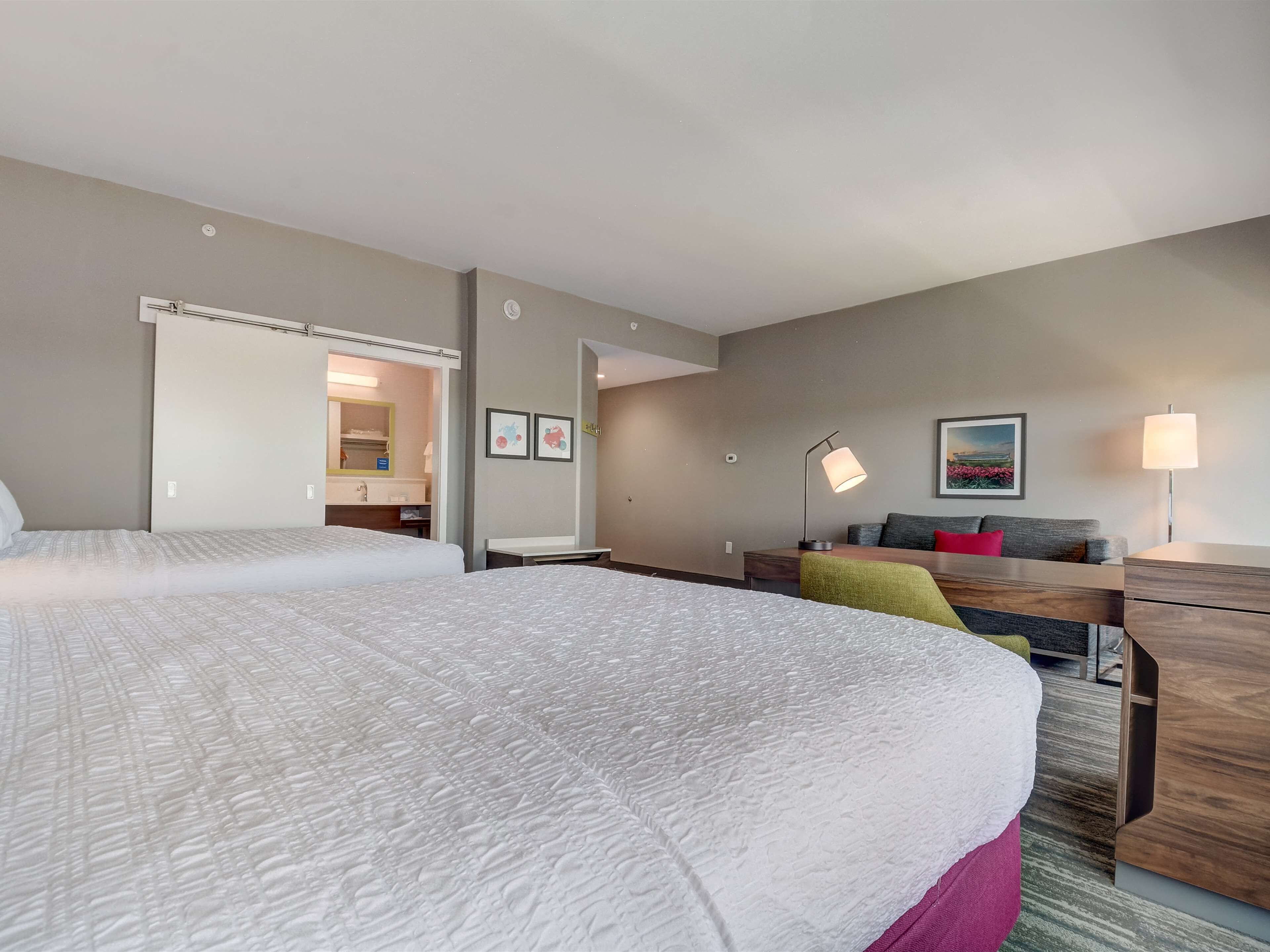 A hotel room with two beds, a desk, a chair, a lamp, and a painting on the wall.