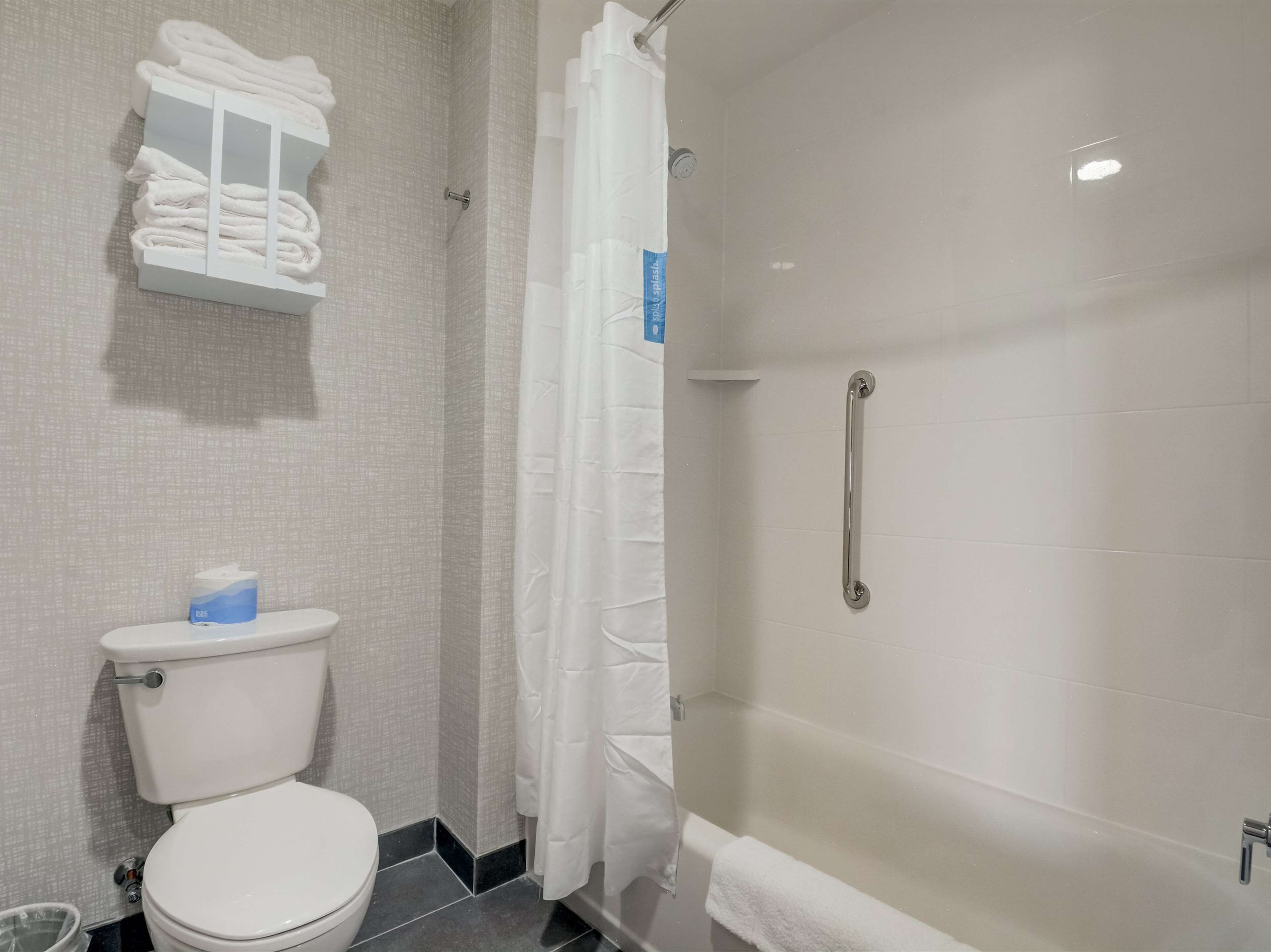 Une salle de bain comprenant des toilettes, une étagère avec des serviettes pliées, une douche avec une porte en verre et une barre d’appui.