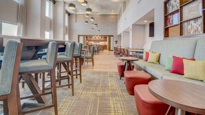 A spacious hotel lobby with modern furniture, a seating area with a sofa and ottomans, and a kitchen visible in the background.