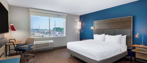A hotel room with a large bed, a desk, a chair, a window with a city view, and a wall-mounted TV.