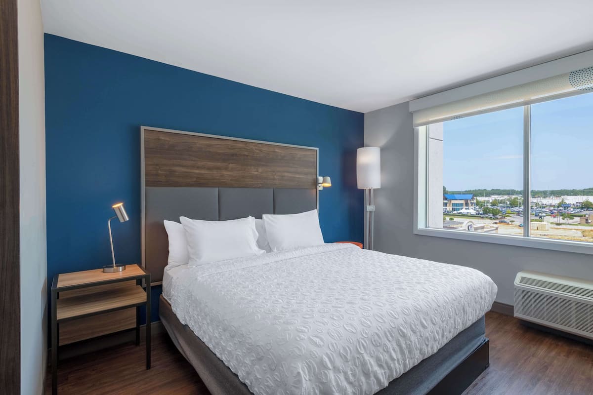 Quarto de hotel moderno com uma cama grande, uma mesa de cabeceira, uma luminária e vista da cidade por uma janela ampla.
