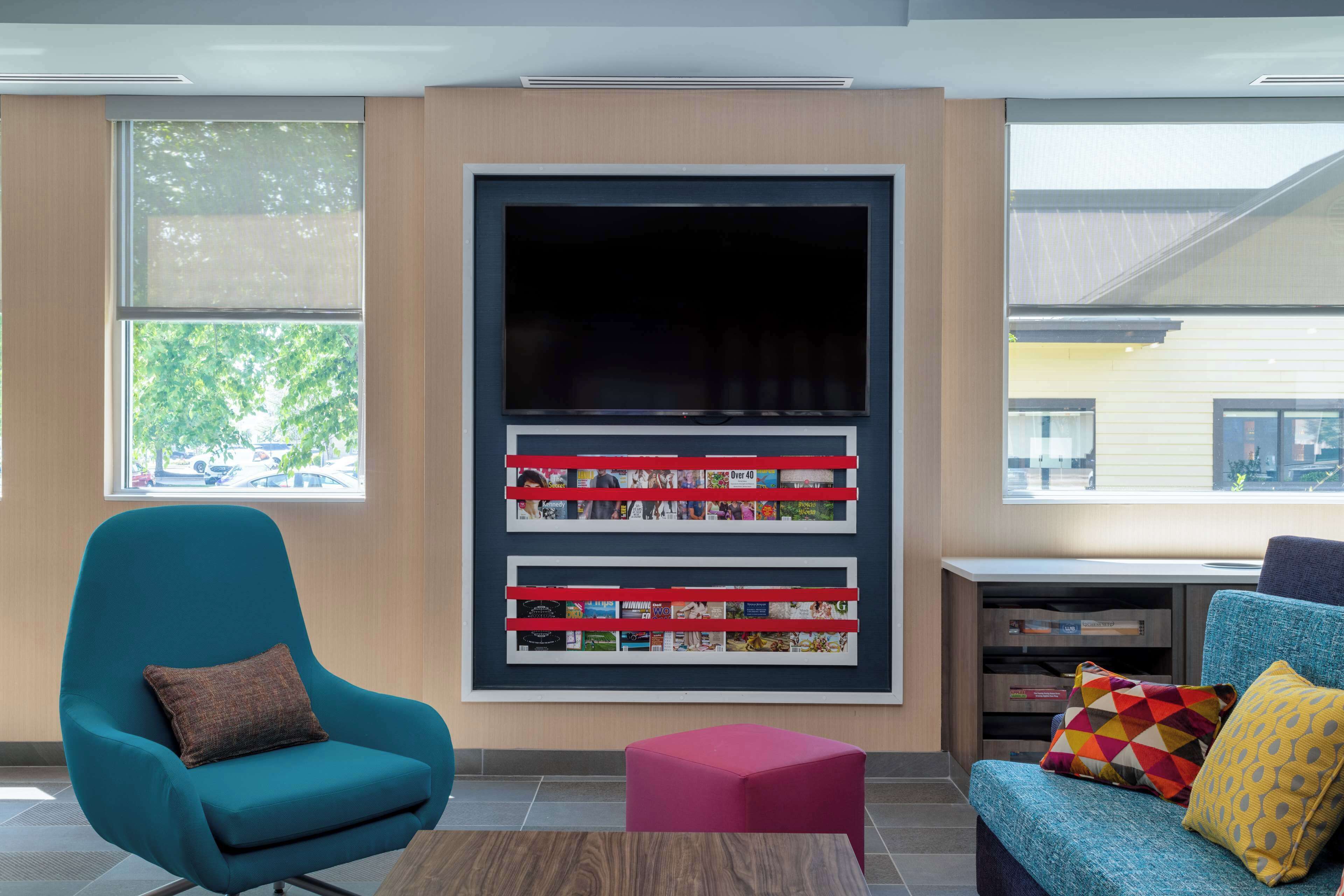 Um lobby moderno com uma poltrona azul-esverdeada, um banquinho vermelho e um sofá azul com almofadas coloridas. Uma grande estante com revistas está fixada na parede.
