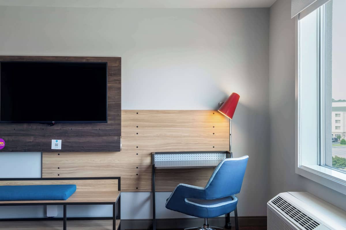 Quarto moderno com televisão na parede, uma cadeira azul, um abajur vermelho e uma janela com vista para edifícios.