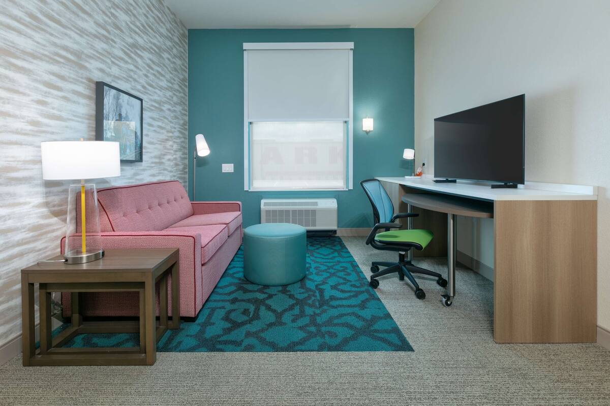 Hotelkamer met een roze bank, een bureau met een televisie, een lamp, een stoel en een kamerkleed met dessin.