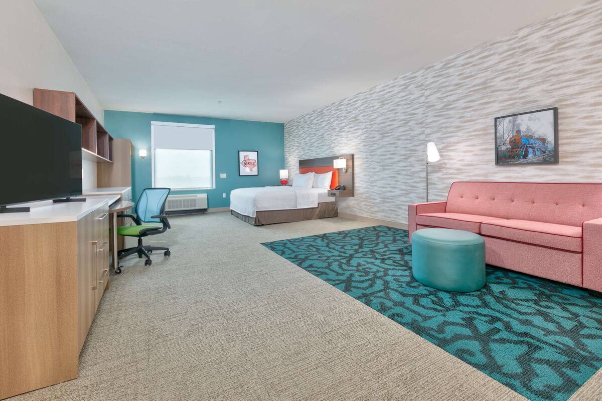 Een hotelkamer met een bed, een bureau, een stoel, een roze bank, een kamerkleed met patroon en een schilderij aan de muur.