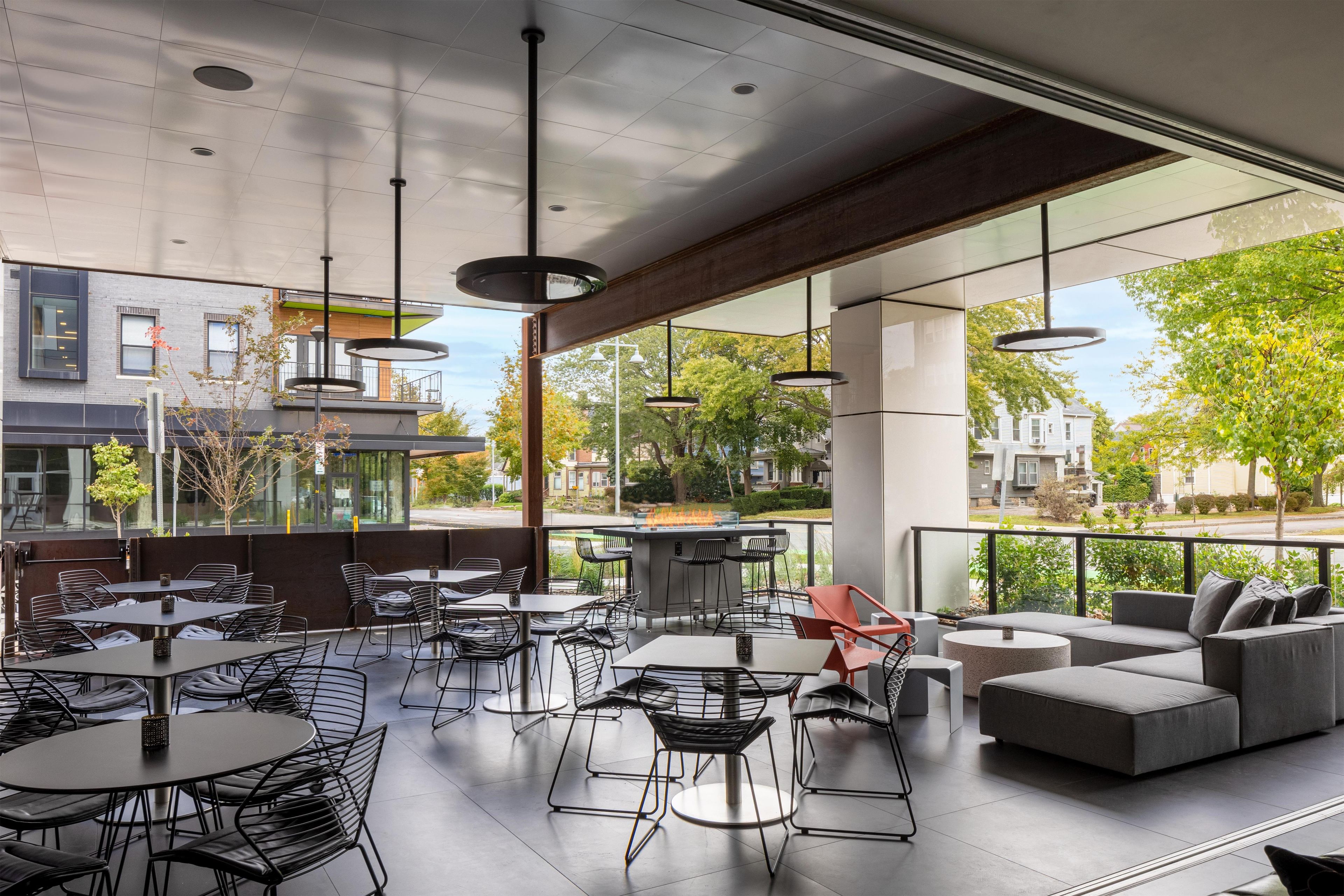 An outdoor seating area with tables and chairs, large windows, and a view of the surrounding neighborhood.