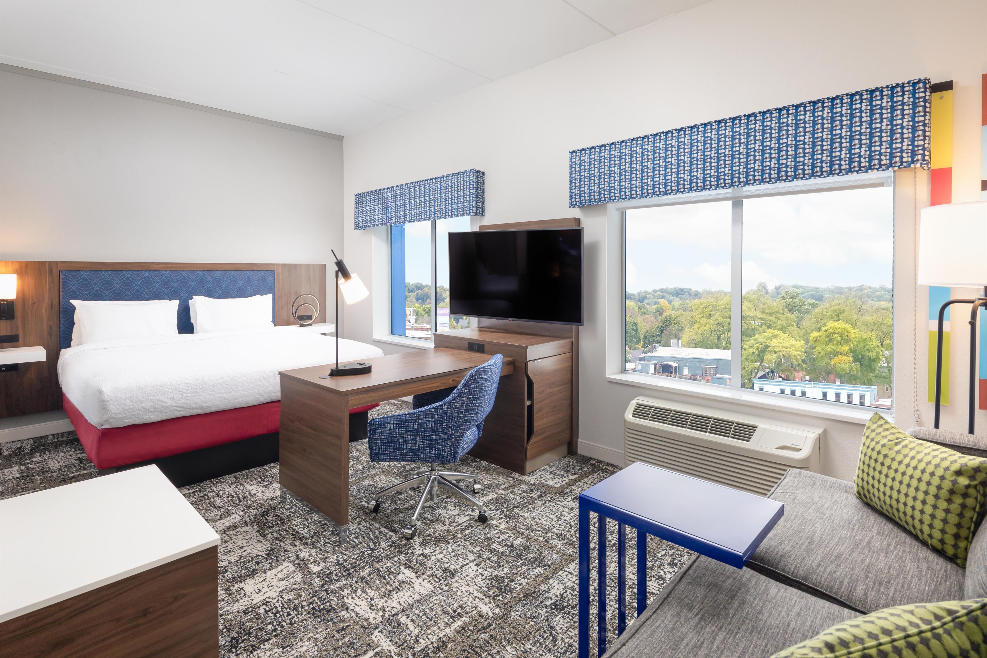 A hotel room with a bed, desk, TV, and a view of the city.