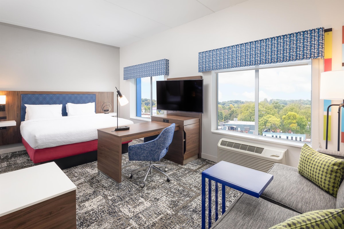 A hotel room with a bed, desk, TV, and a view of the city.