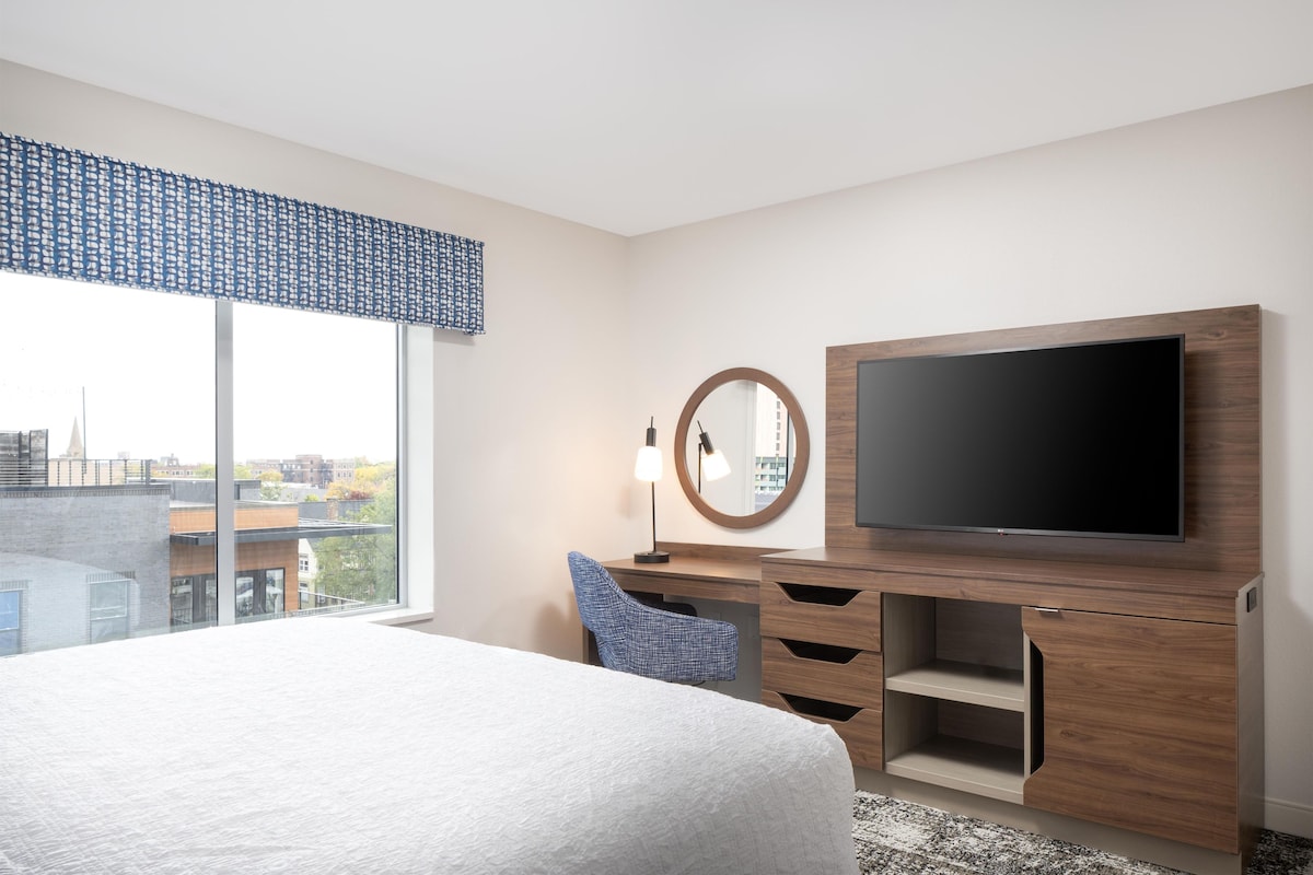 A hotel room with a large bed, a flat-screen TV mounted on a wooden cabinet, a desk with a chair, and a window with a view of buildings.