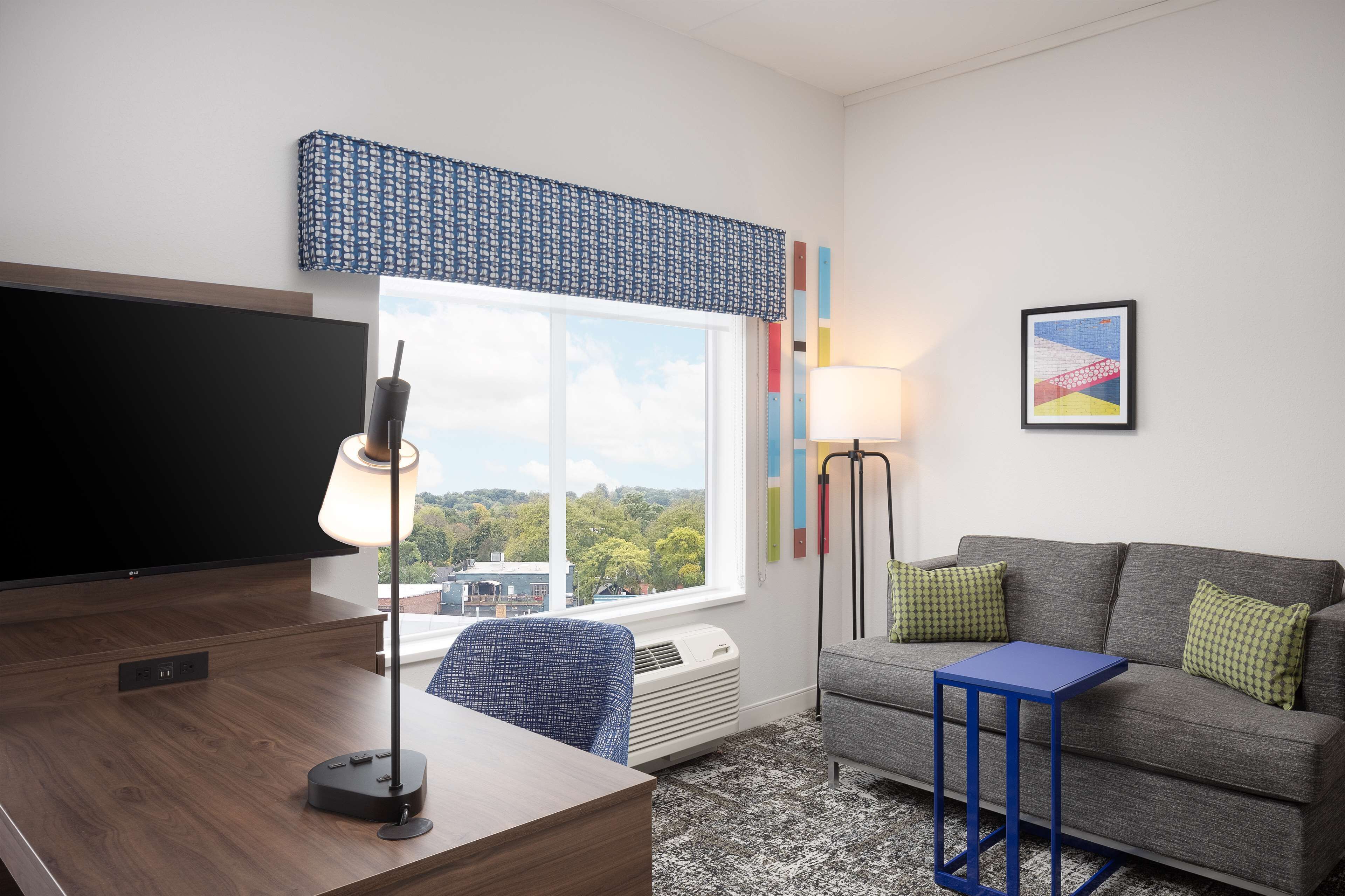 A modern living room with a flat-screen TV, a desk, a blue lamp, a grey sofa with green cushions, and a large window with a view of trees and buildings.