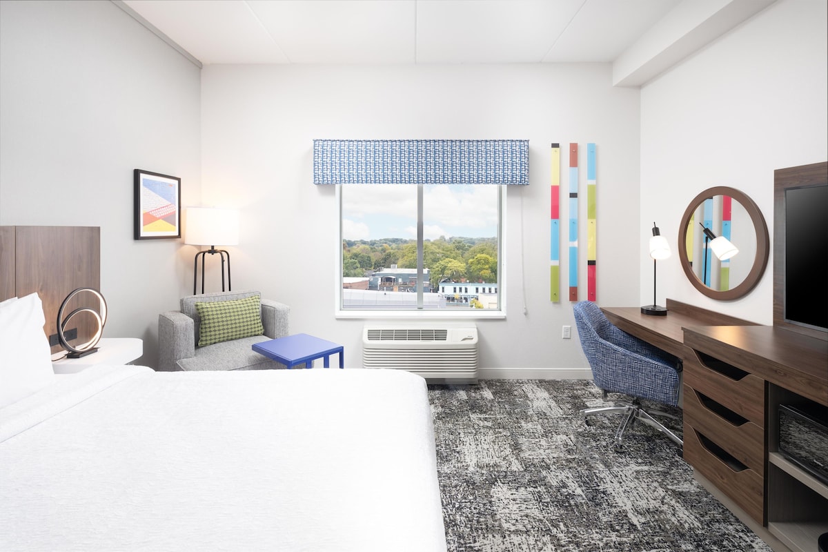 A hotel room with a bed, a desk, a chair, a TV, a window with a view, and a radiator.