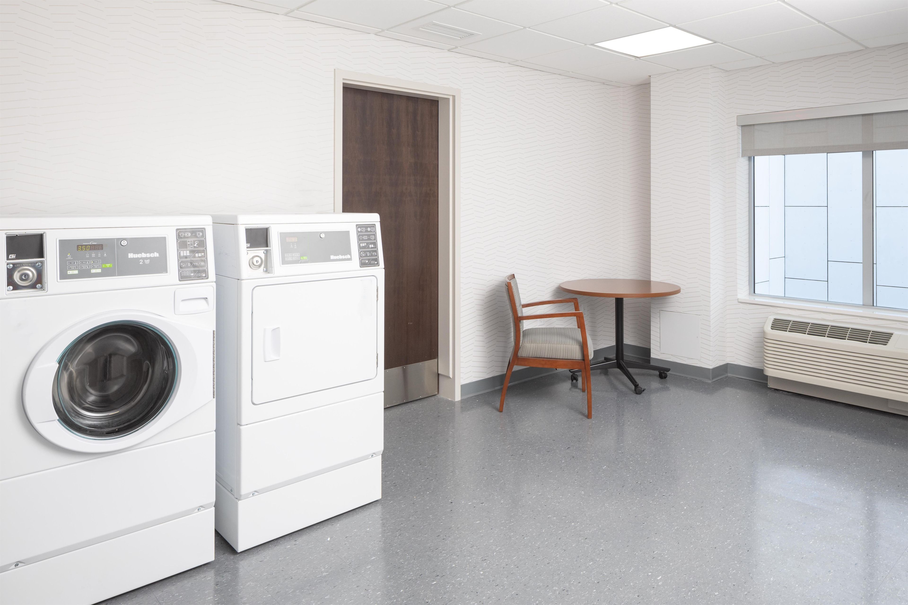 A laundry room with two front-loading washing machines, a wooden chair, a small table, and a window with blinds.