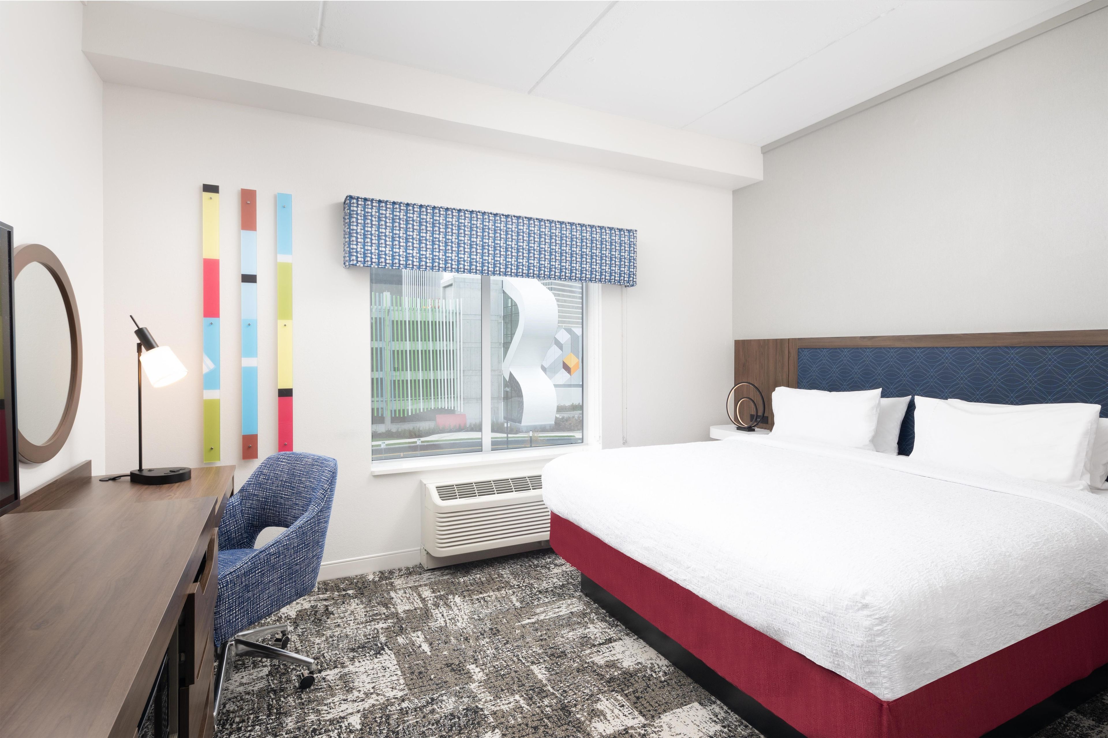 Habitación de hotel con cama, escritorio, silla, ventana con vistas y alfombra estampada.
