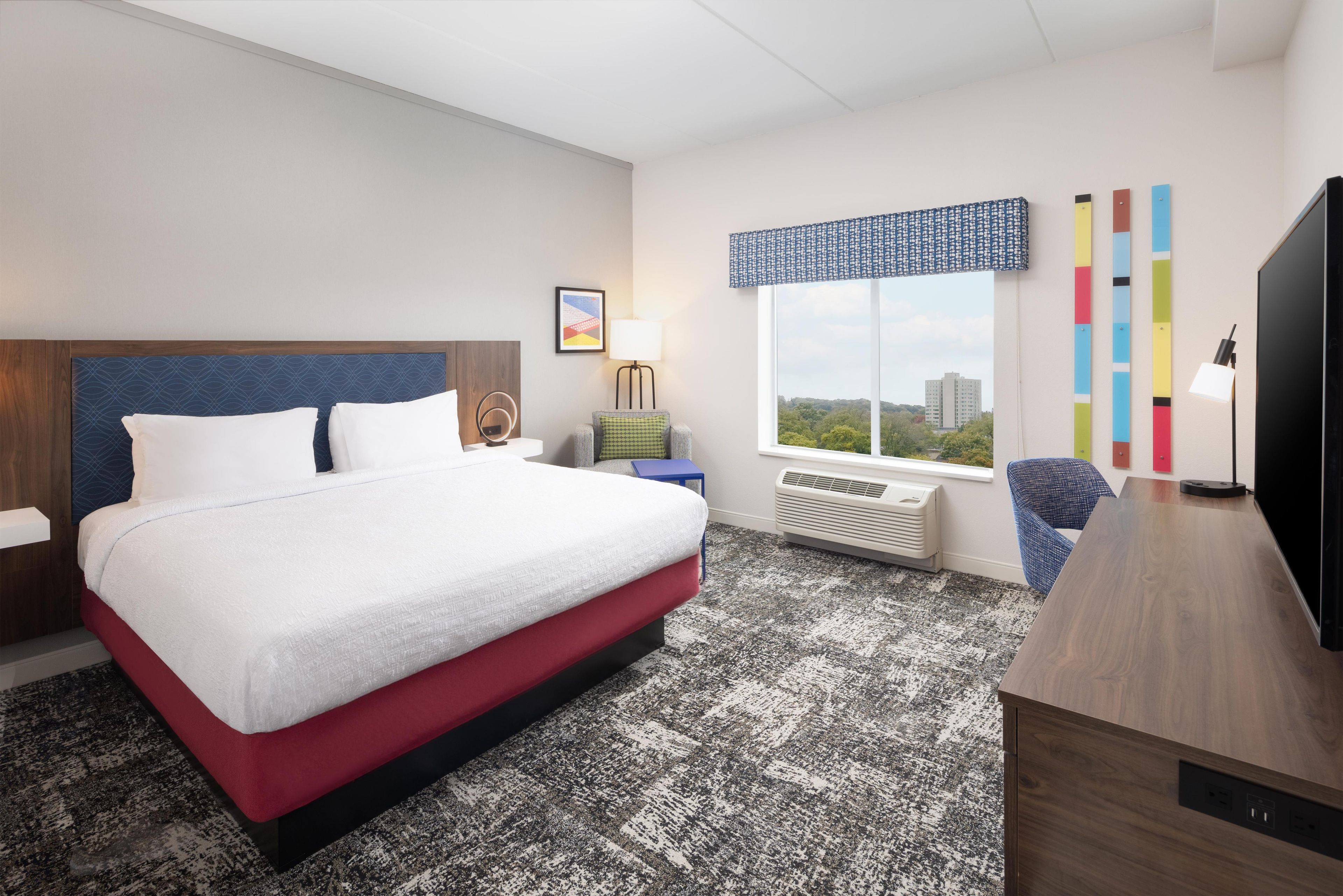 A hotel room with a bed, a desk, a chair, a window with a view, and a wall with a colorful panel.