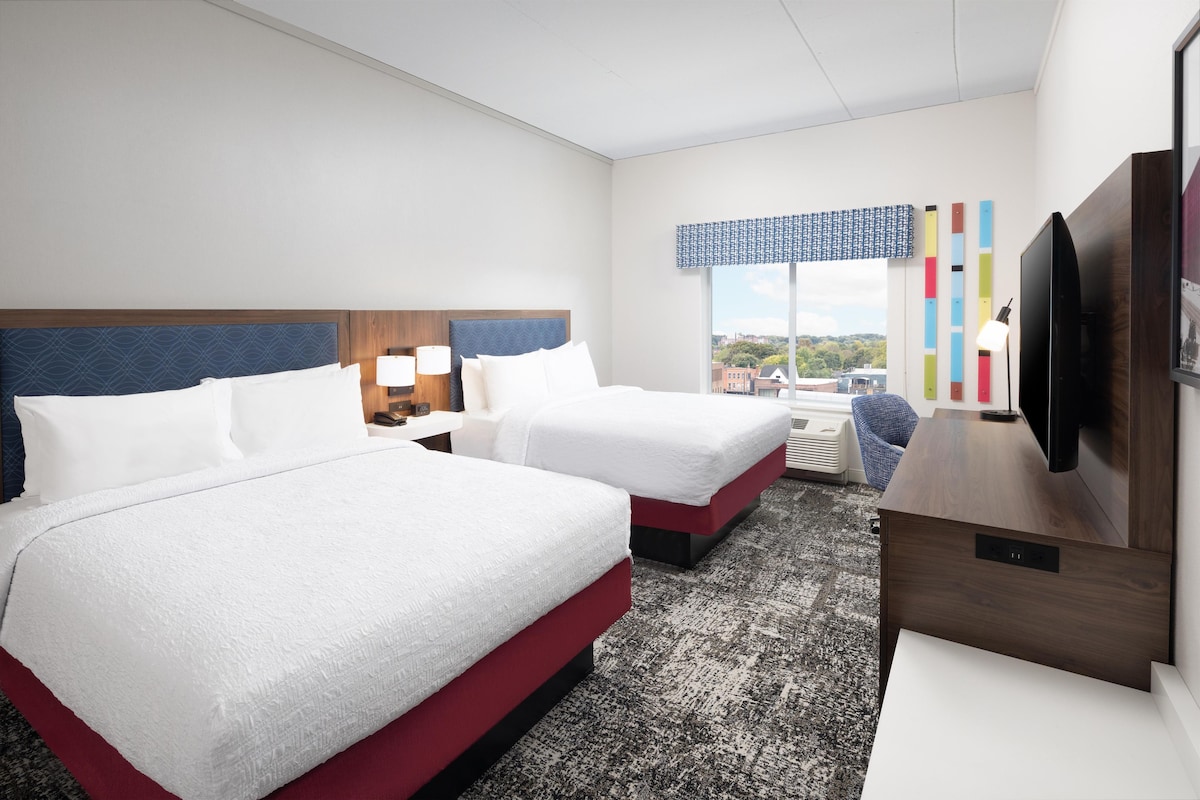 A hotel room with two beds, a TV, a desk, and a window with a city view.