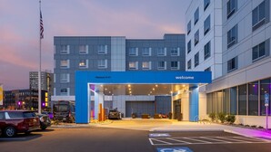 En moderne hotelindgang med et blåt skilt, der siger "Velkommen", et parkeringsområde med parkeringspladser for handicappede, og et flagstang med et amerikansk flag.