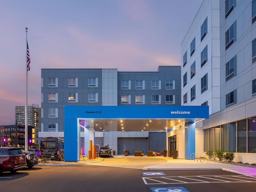 A modern hotel entrance with a blue sign reading "Welcome," a parking area with handicapped parking, and a flagpole with an American flag.