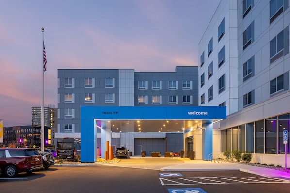A modern hotel entrance with a blue sign reading "Welcome," a parking area with handicapped parking, and a flagpole with an American flag.