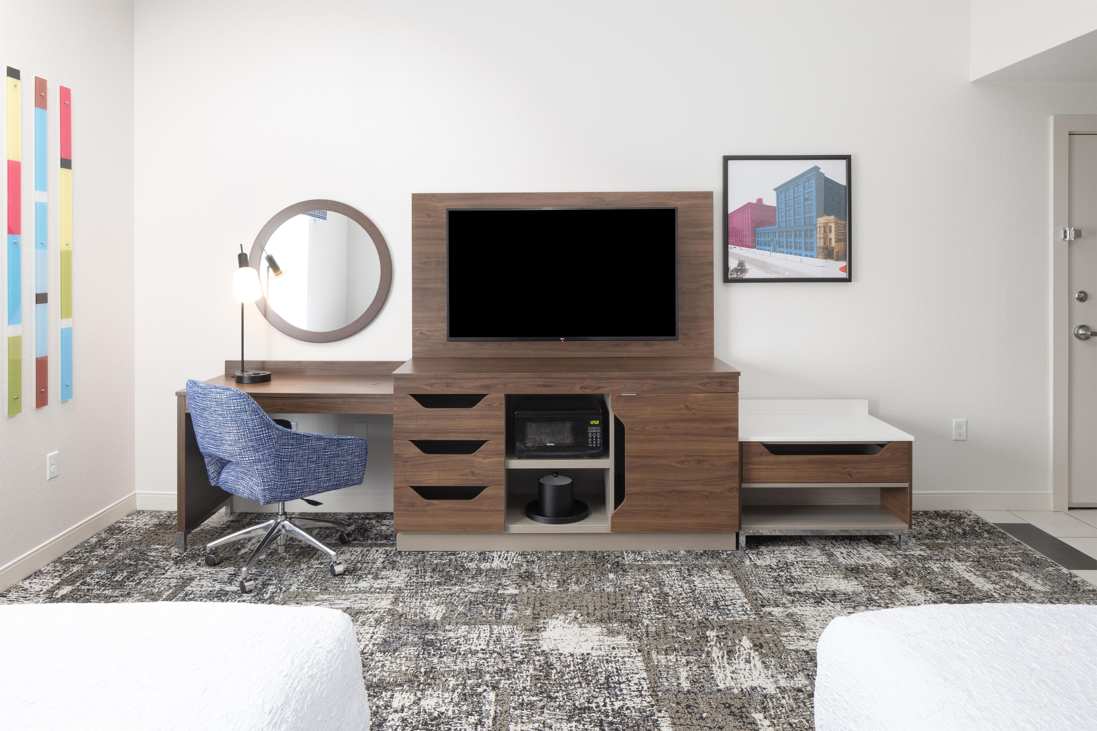 A hotel room with a wooden desk, a flat-screen TV, a blue chair, a round mirror, a microwave, and a framed cityscape picture on the wall.