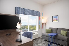 A modern living room with a flat-screen TV, a desk, a standing lamp, a sofa, and a window with a view of greenery. - Hampton Inn & Suites Rochester Downtown (Rochester)