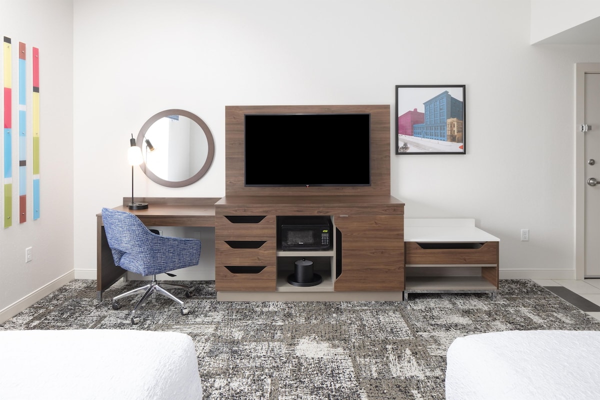 A hotel room with a wooden desk, a flat-screen TV, a chair, a microwave, and a framed picture on the wall.