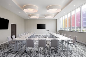 A conference room with a long table, chairs, a flat-screen TV, and large windows with a view of colorful buildings.