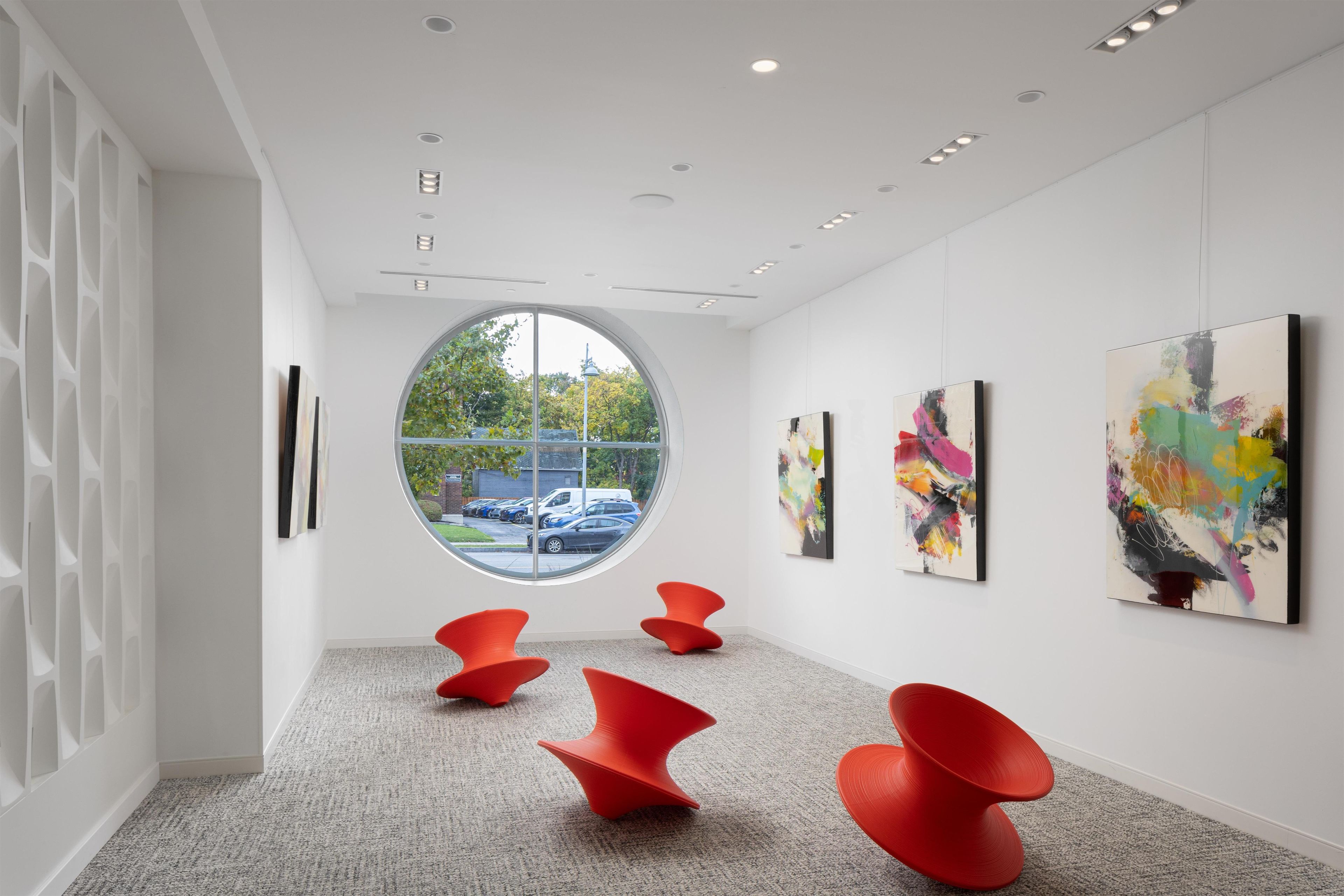 Una galería de arte moderno con un ventanal circular central, sillas rojas y pinturas abstractas en las paredes.