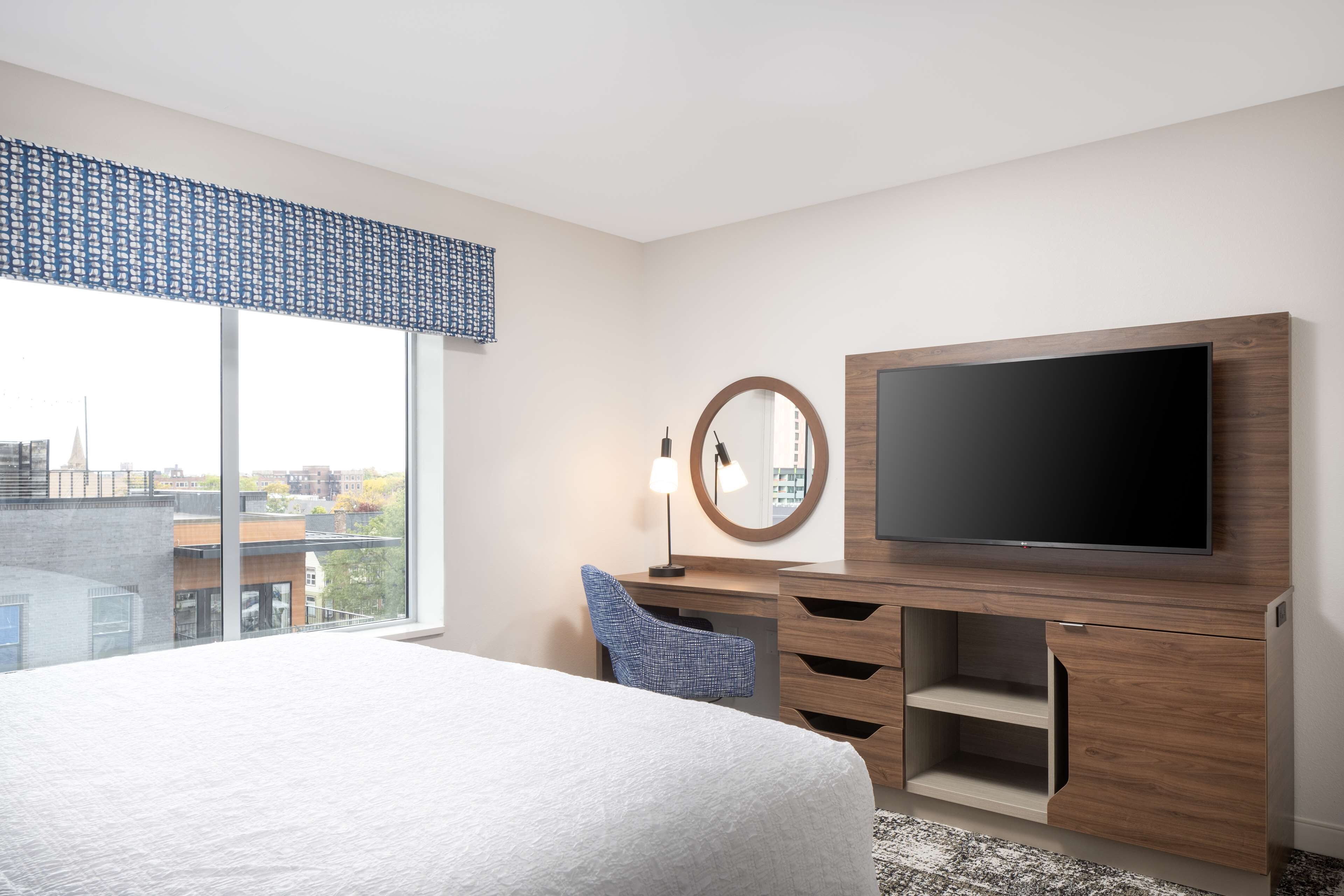 A modern hotel room with a large bed, a flat-screen TV, a desk with drawers, a chair, and a large window offering a city view.
