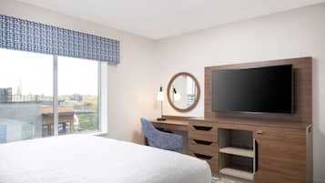 A modern hotel room with a large bed, a flat-screen TV, a desk with drawers, a chair, and a large window offering a city view.