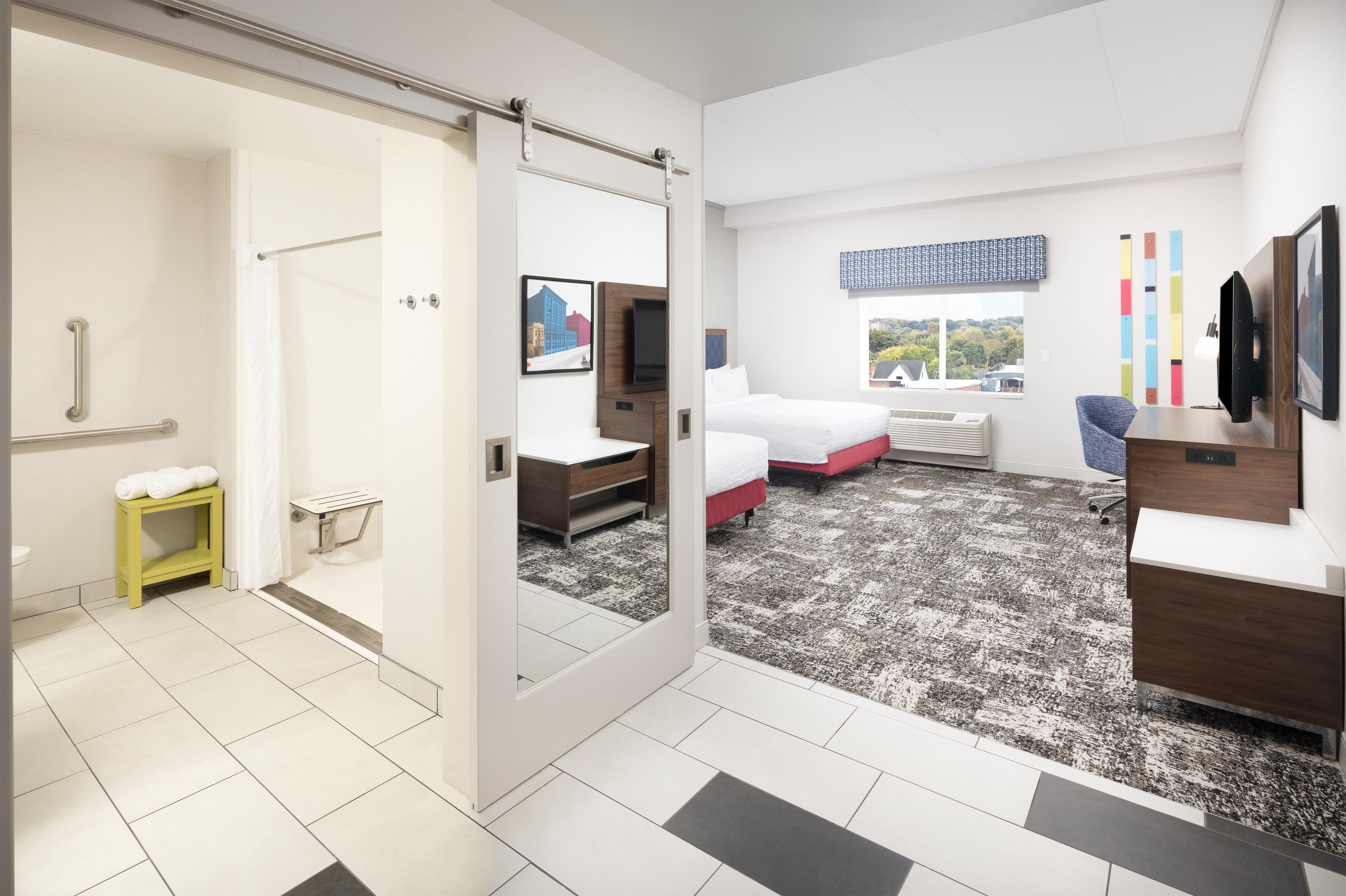 A modern hotel room with a sliding door, a bed, a desk, and a window with a city view.