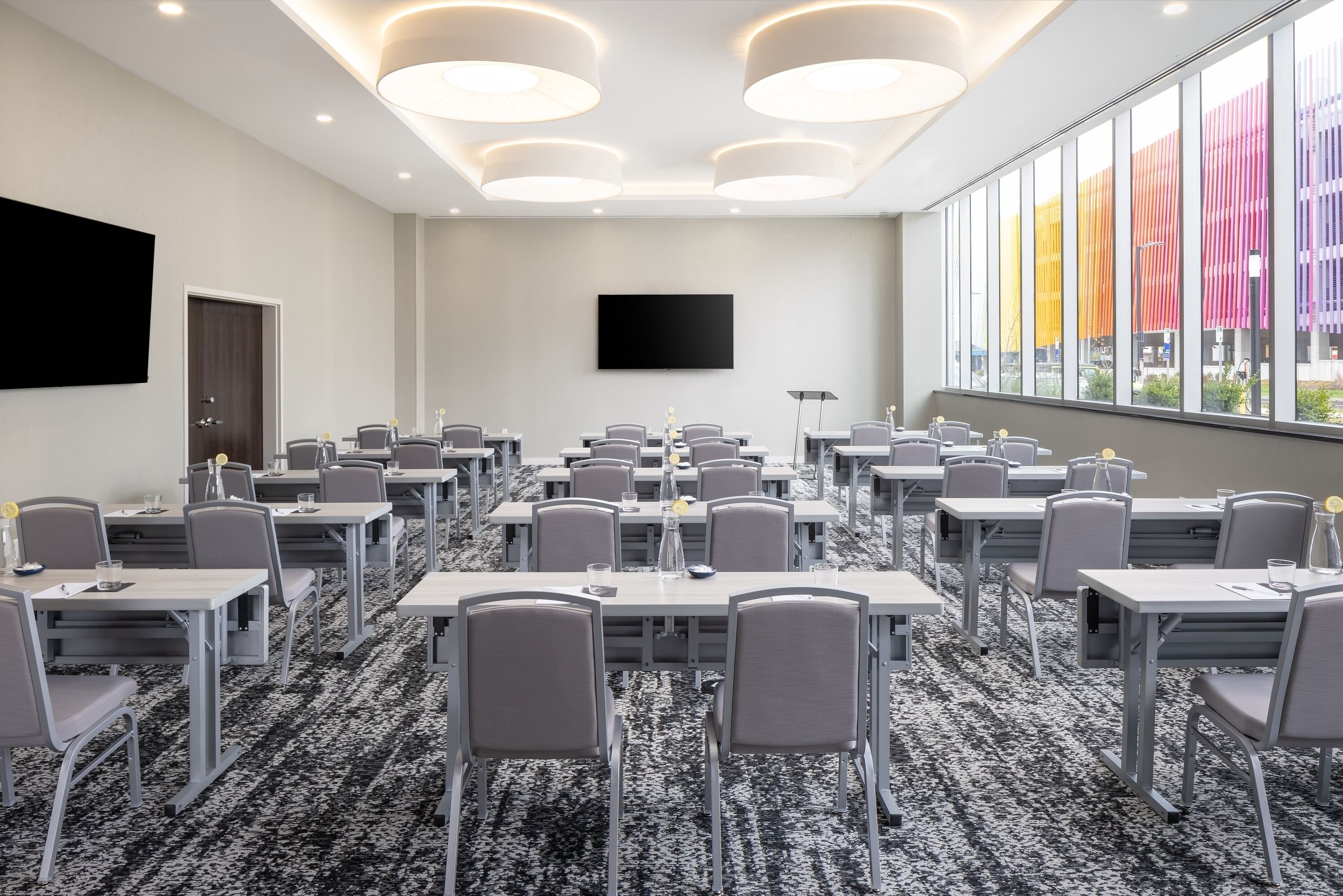 A conference room with multiple tables and chairs, a large flat-screen TV, and a view of colorful buildings through large windows.