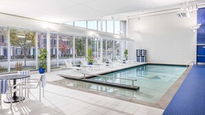 A modern indoor swimming pool with clear glass walls, lounge chairs, and a table with chairs.