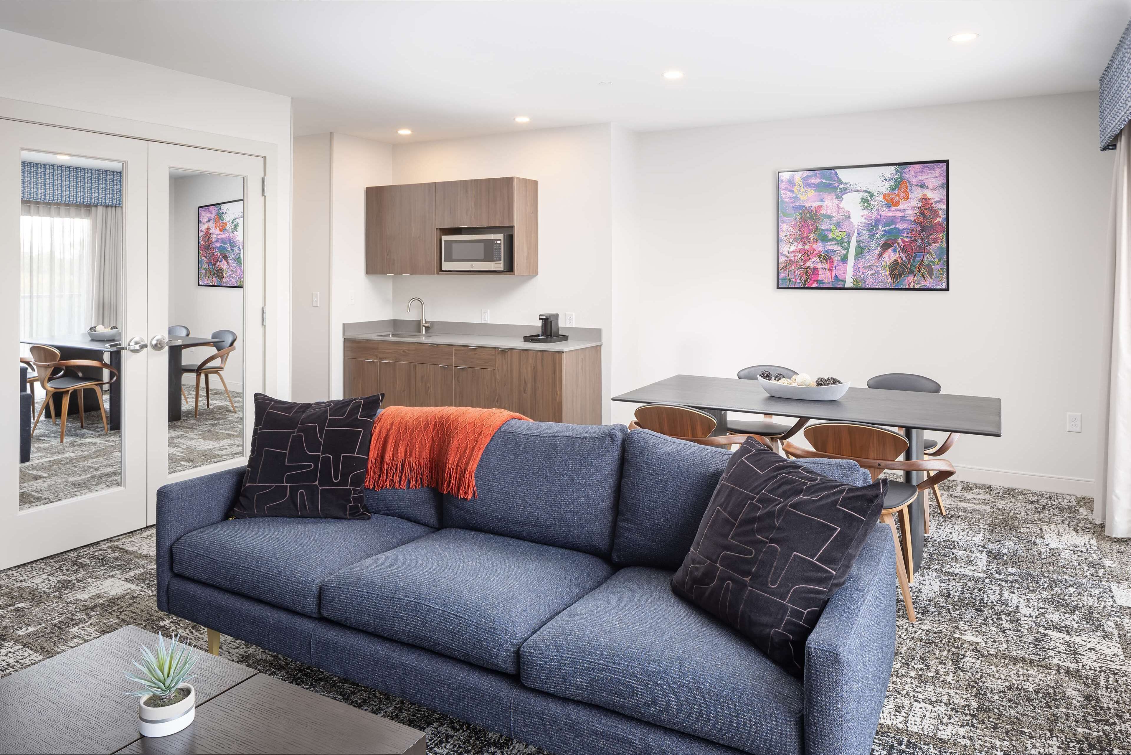 Una sala de estar moderna con un sofá azul, una mesa de centro con una planta, una zona de comedor con una mesa y sillas, una cocina con microondas y obras de arte en las paredes.
