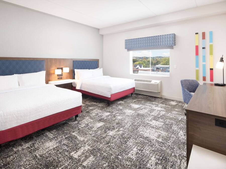 A hotel room with two beds, a desk, a chair, a TV, a window with a view, and a wall with colorful decor.