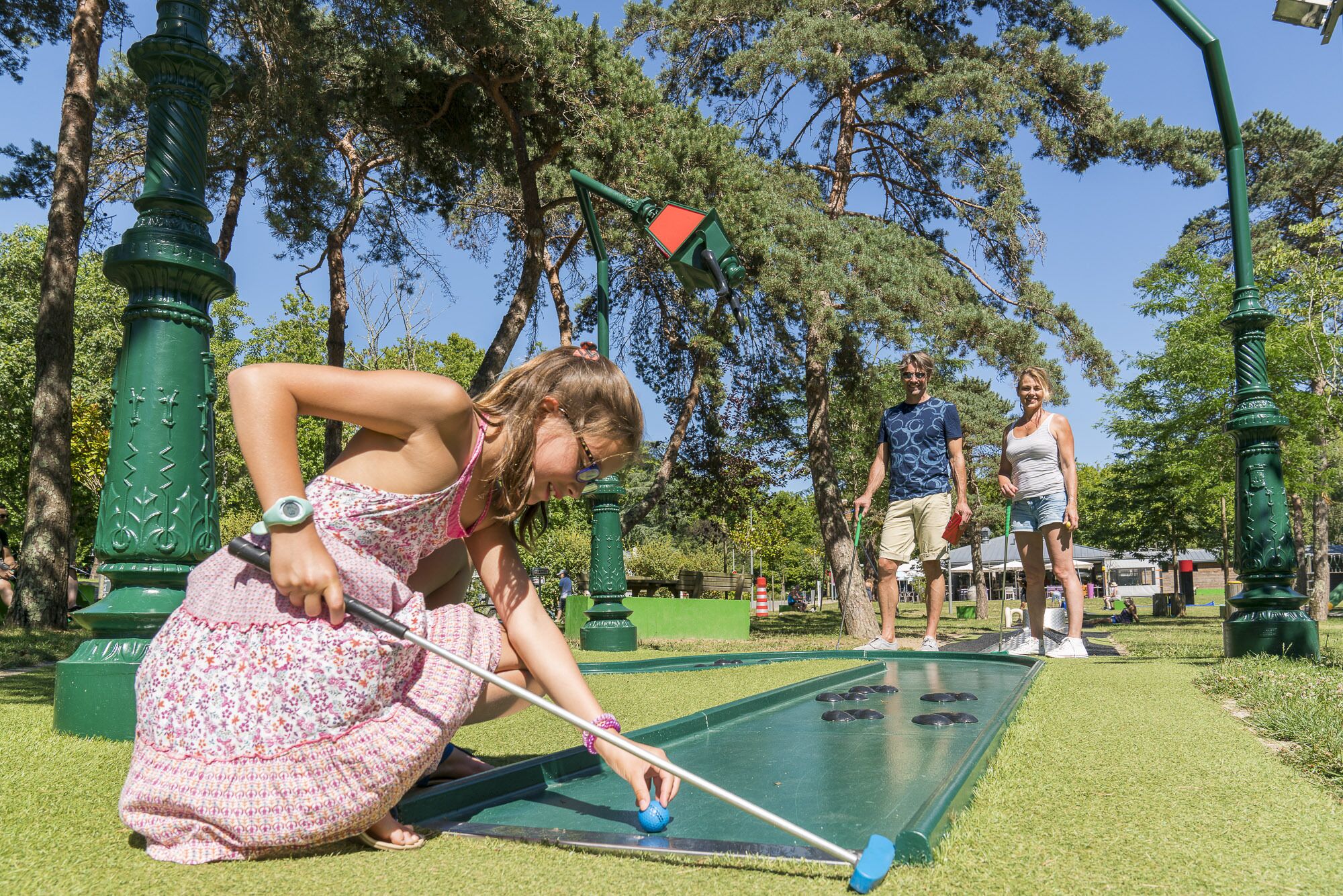 A person playing shuffleboard on a green court with a backdrop of trees and a clear sky.