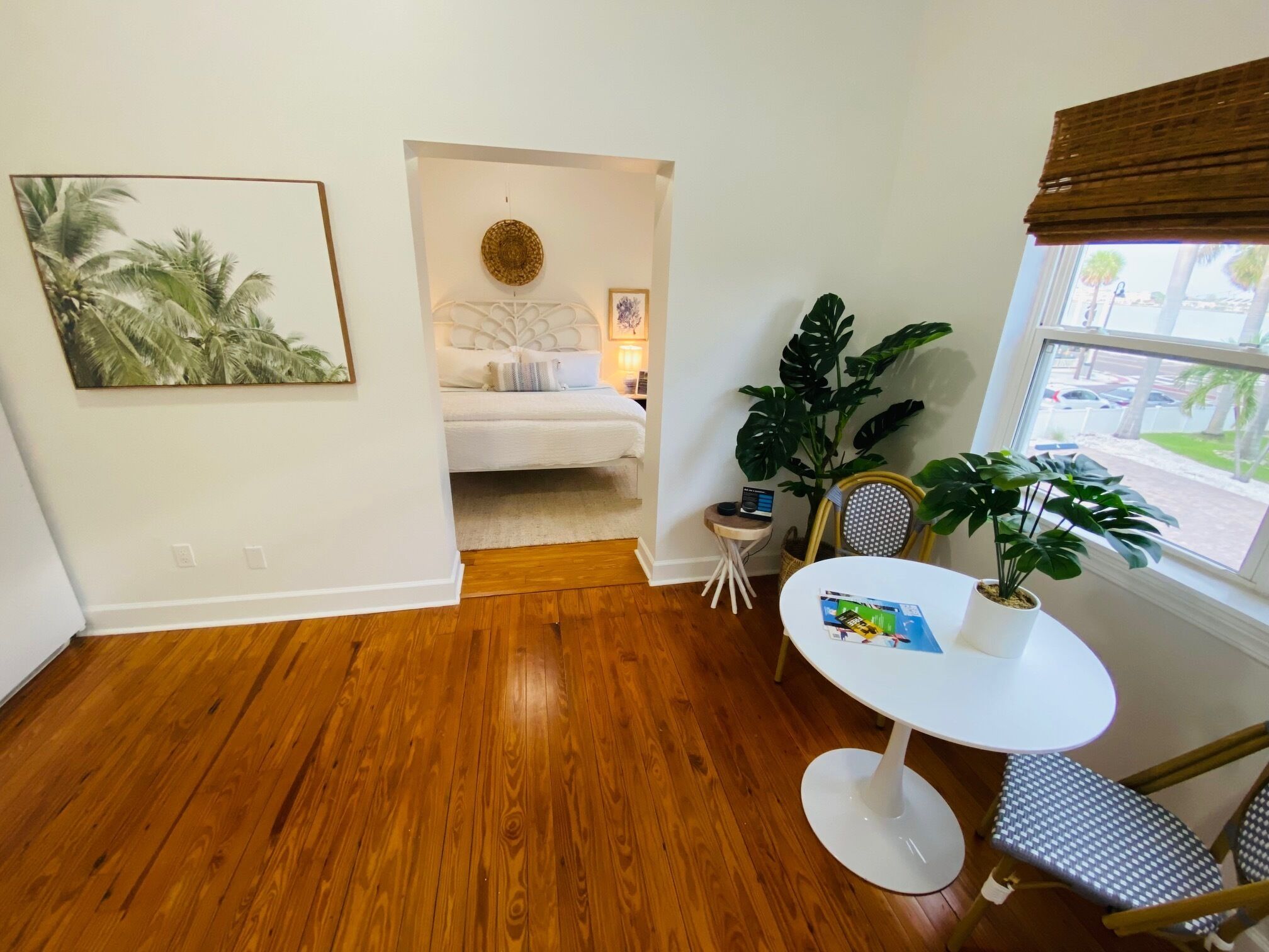 Una habitación con piso de madera, una cama con ropa de cama blanca, una pequeña mesa con una planta y una ventana con vistas a palmeras.